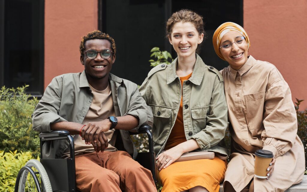 Group of positive young multi-ethnic friends sitting on bench and drinking coffee with handicapped man outdoors