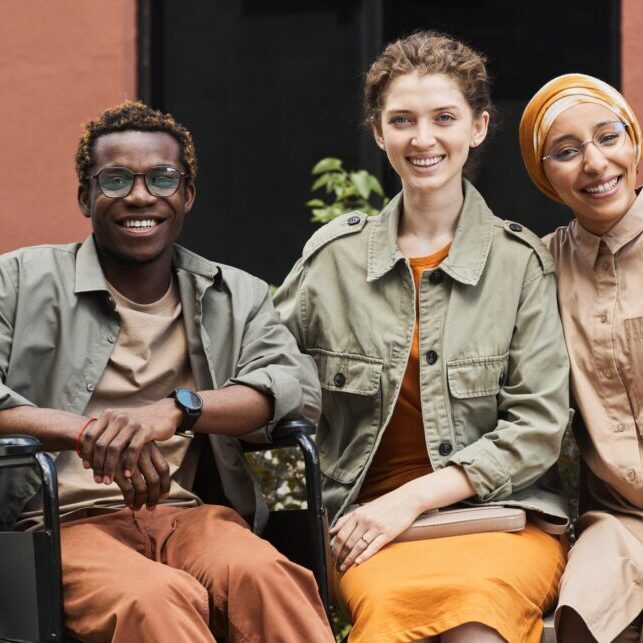 Group of multi-ethnic friends outdoors Group of positive young multi-ethnic friends sitting on bench and drinking coffee with handicapped man outdoors