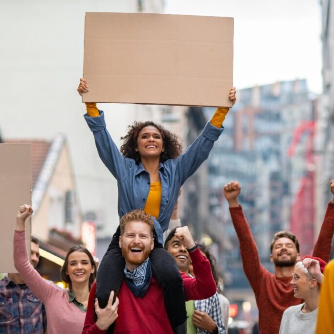 Happy group of people holding blank banner during strike Group of multiethnic people on city street holding blank cardboard placard celebrating victory during a protest. Young group of content men and smiling women marching through a city. People protesting on road with empty sign.